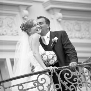 Ein glückliches Hochzeitspaar. Die Braut lächelt. Der Bräutigam küsst sie. Beide haben die Hände auf einem Geländer. Darauf sieht man die Trauringe.